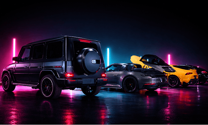 Row of luxury cars at night bathed in neon pink and blue lights, including a black Mercedes G‑Wagon, a gray sports car, and a yellow Lamborghini-like supercar on a glossy surface.